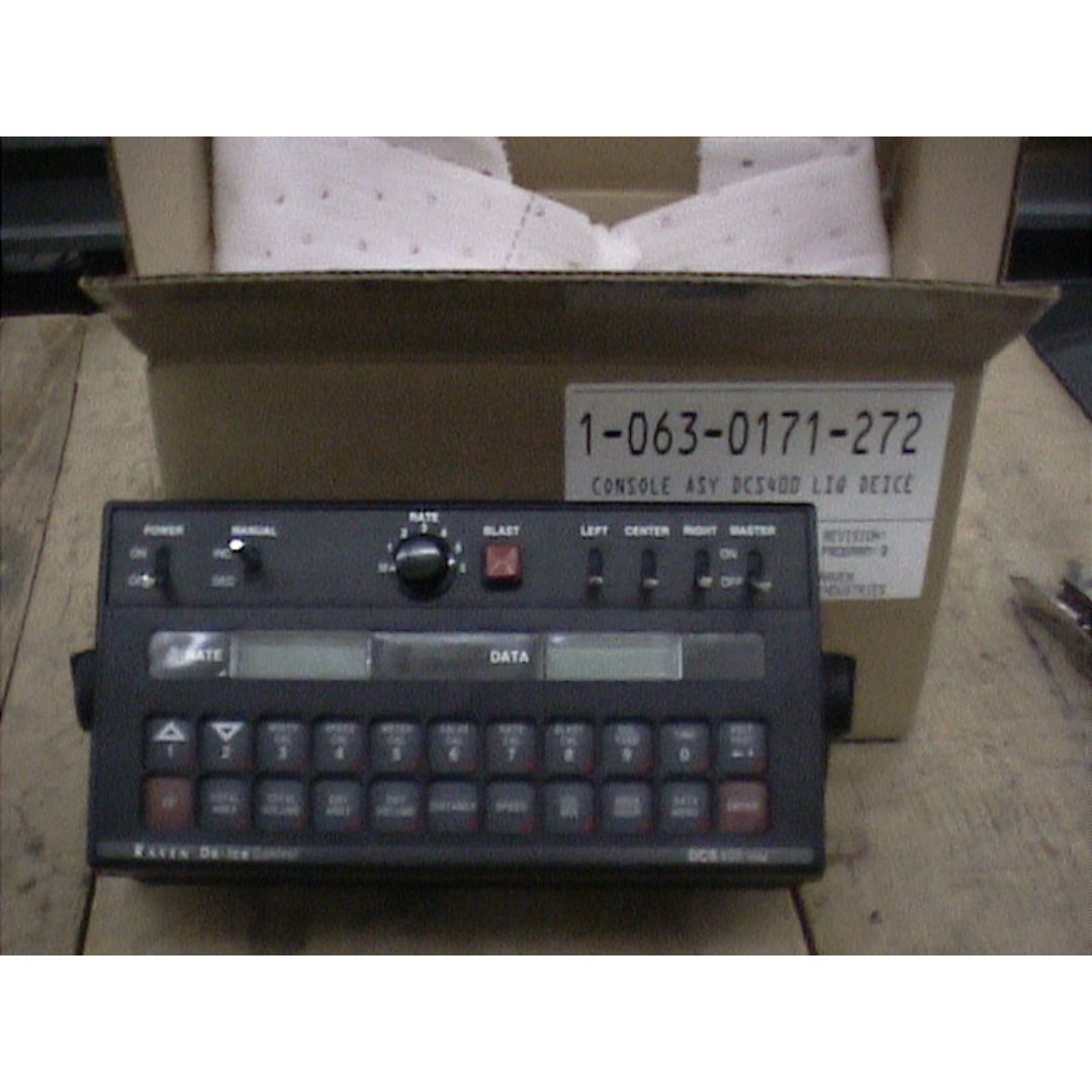 The Raven DCS 400 Liquid De-Icer Console, a black electronic device with multiple buttons and a small screen designed for granular spreader control, sits in front of an open cardboard box on a wooden surface.