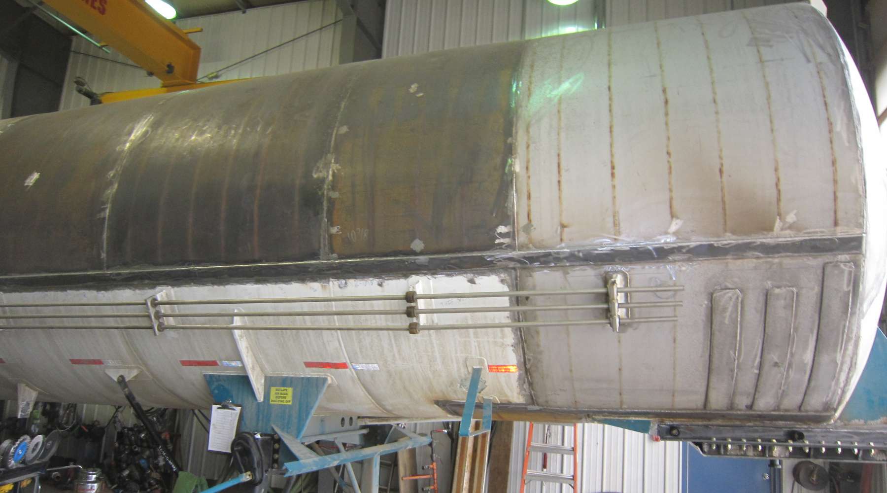 Large cylindrical tank with visible rust and repair patches, supported on blue stilts in an industrial setting.