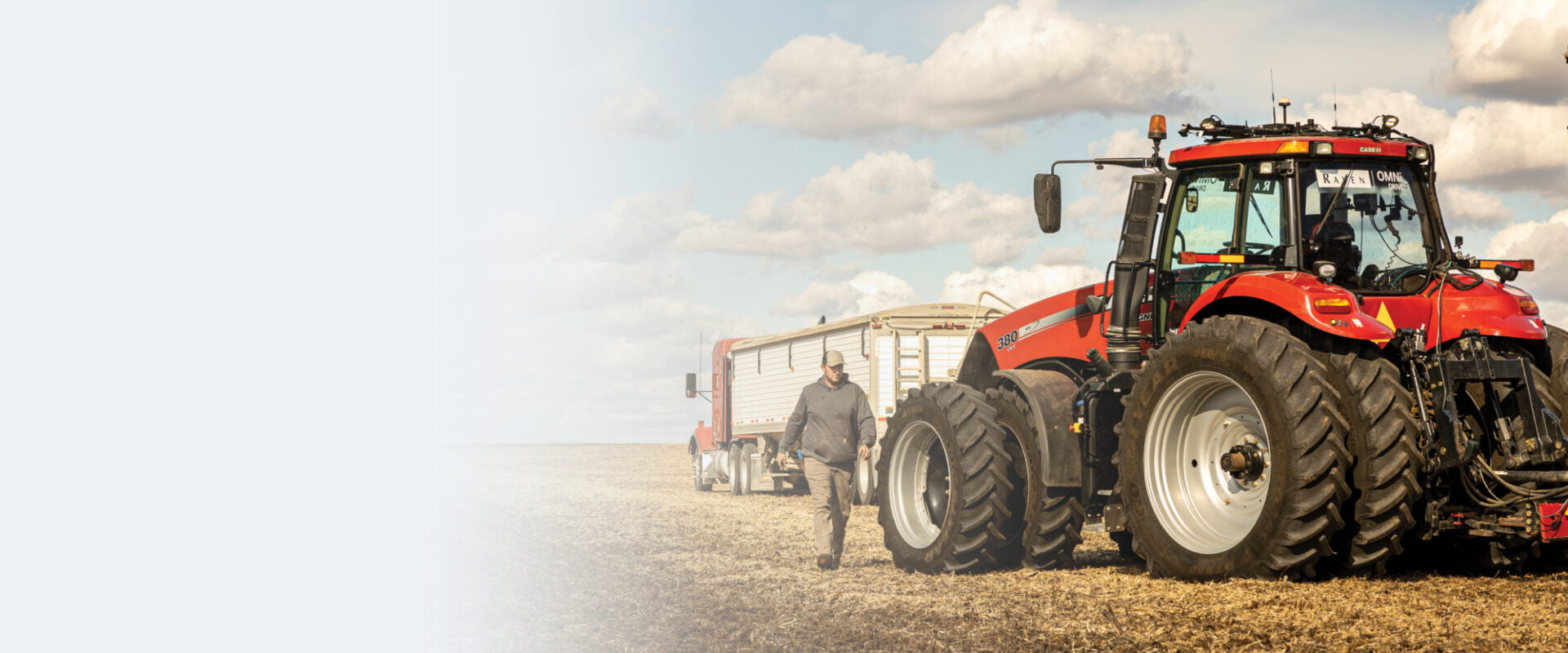 A person walking near a red tractor and a trailer in a vast field under a cloudy sky.