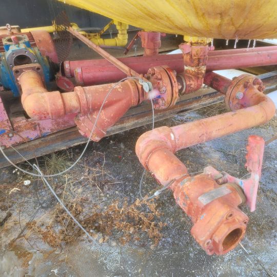Rusted red industrial pipes and fittings attached to a yellow storage tank, with cables and connections visible on a frosty ground.