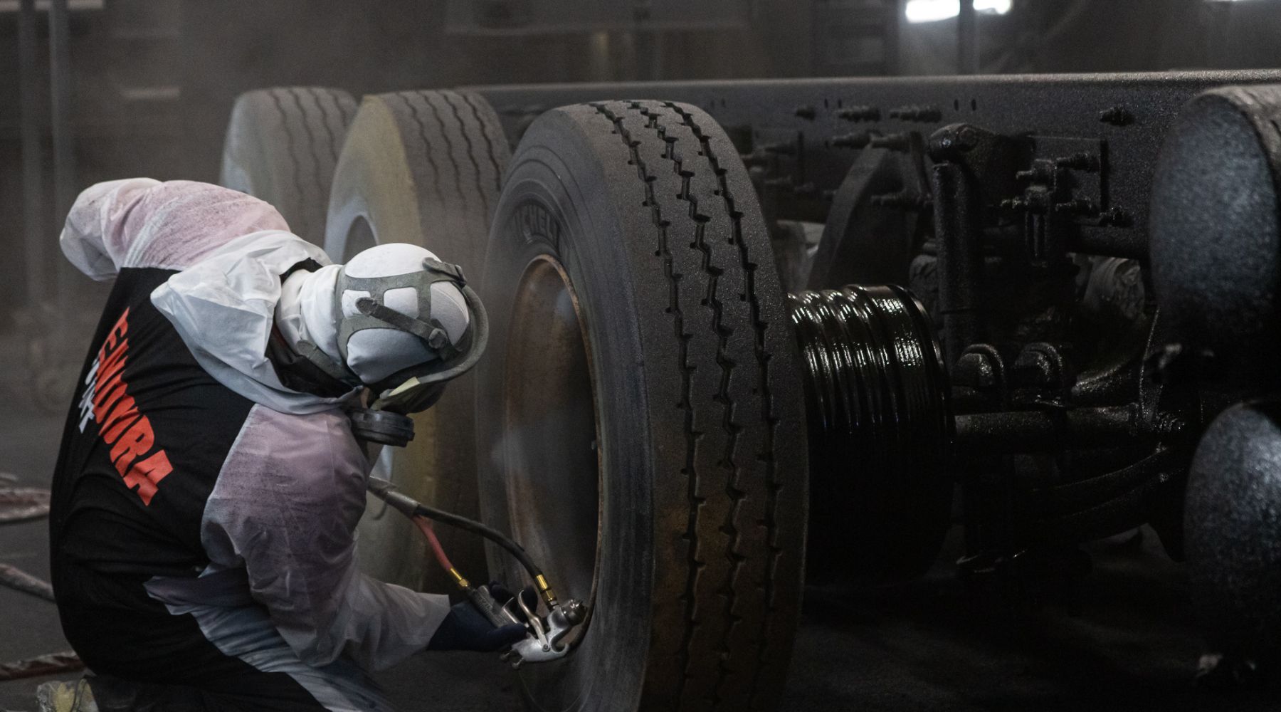 A worker wearing protective gear sprays coating on large truck tires in a dimly lit industrial setting.