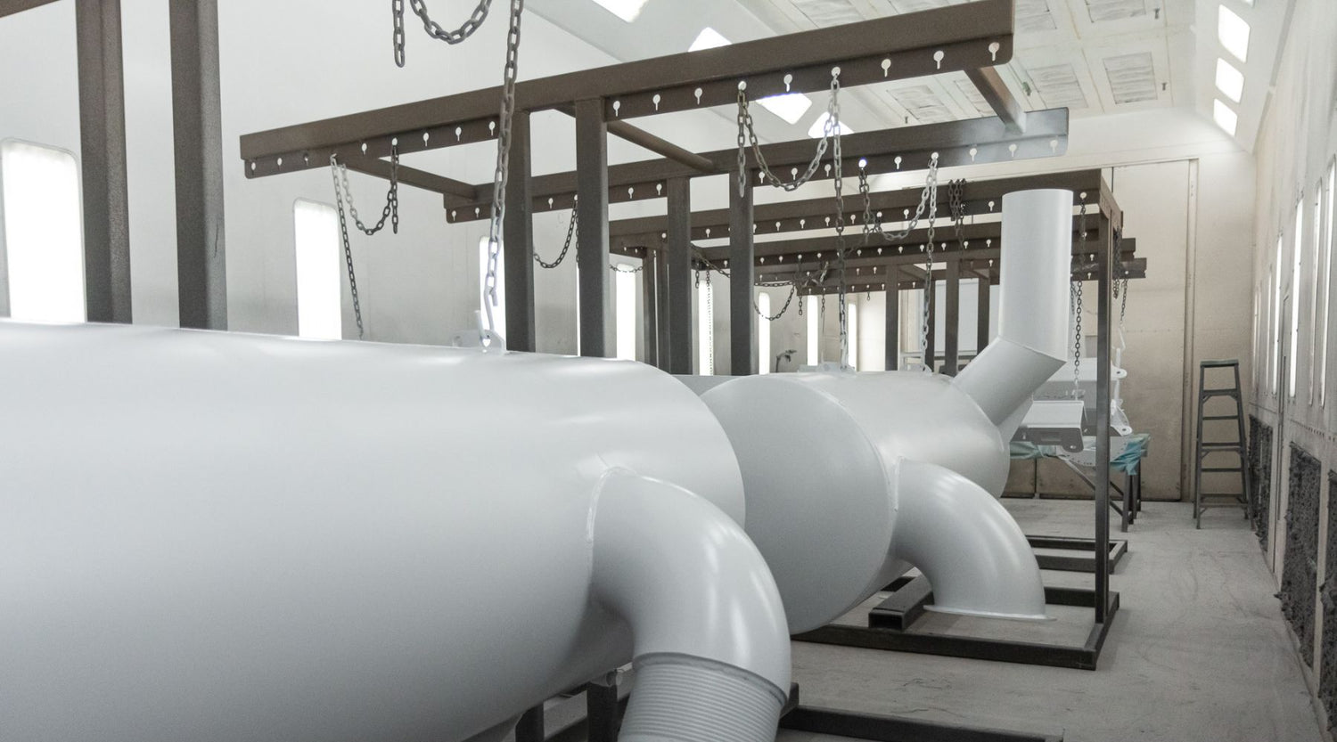Large white industrial components are suspended indoors by chains attached to metal frames.