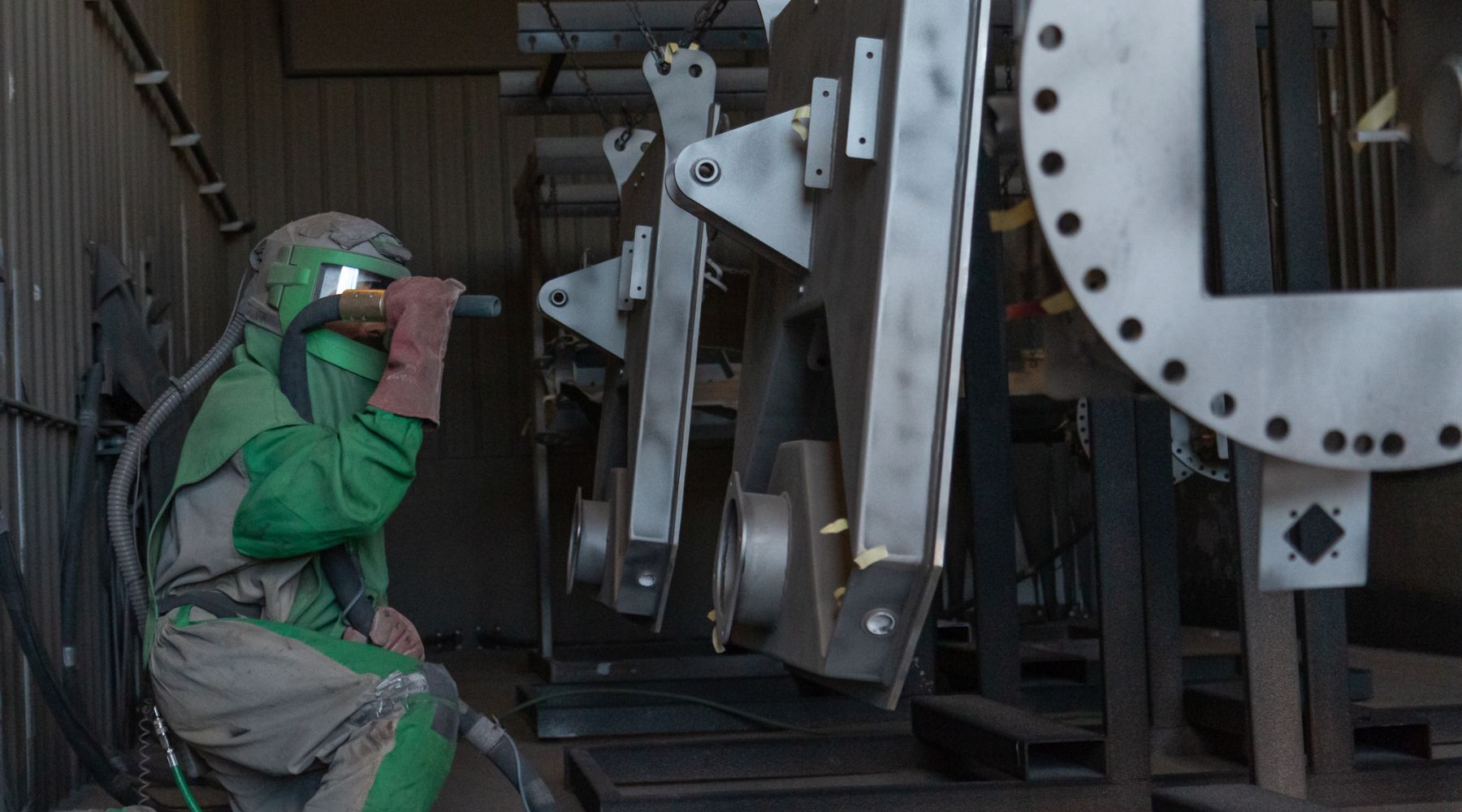 A worker in protective gear operates a sandblasting tool on metal structures in an industrial setting.