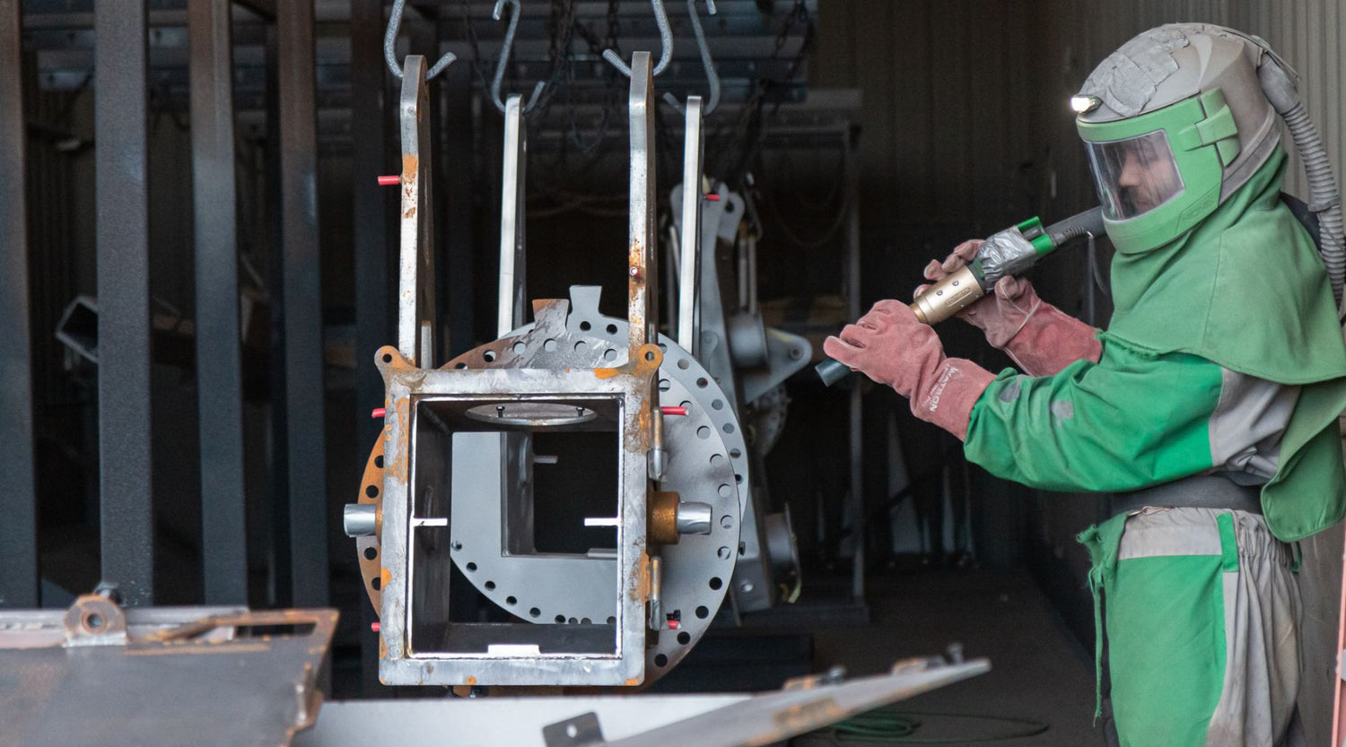 A worker in protective gear sandblasts a metal cylinder in an industrial setting.