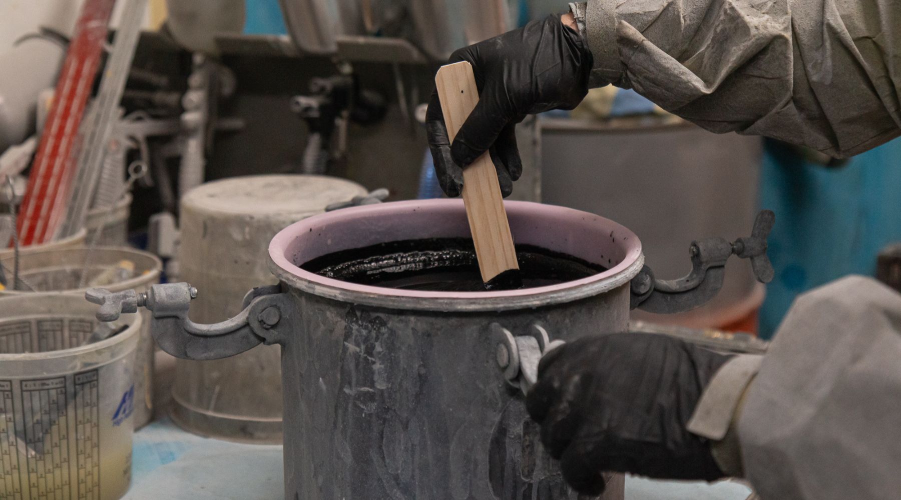 Person in protective gear stirring black liquid in a large industrial container using a wooden stick.