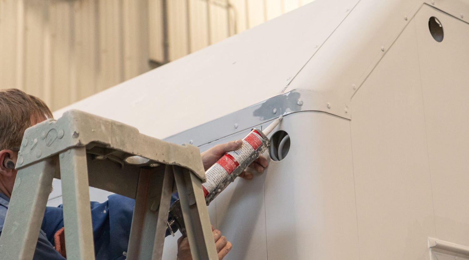 Person on a ladder applying sealant to the corner of a large white structure in an industrial setting.