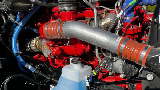 Close-up of a Cummins X15N natural gas engine with various pipes and hoses, featuring a prominent silver tube and a blue fluid container in the foreground.