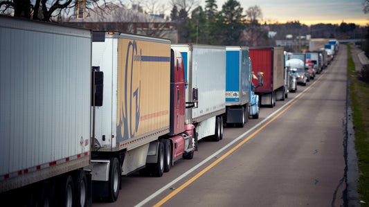 A long line of semi-trucks is parked along the highway, with a cloudy sky and trees in the background.