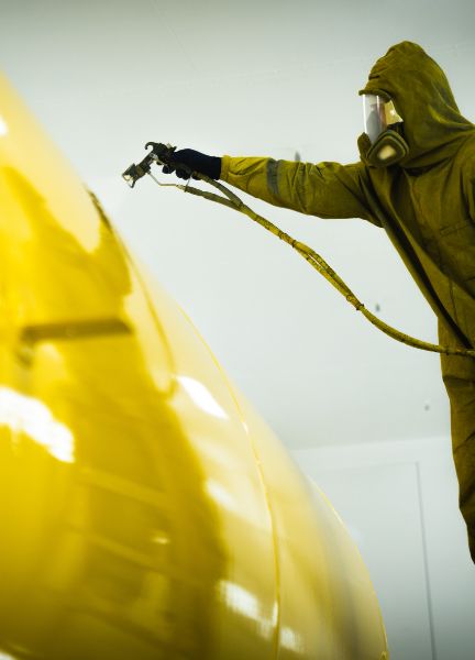 Person in protective gear spray painting a large object in bright yellow.