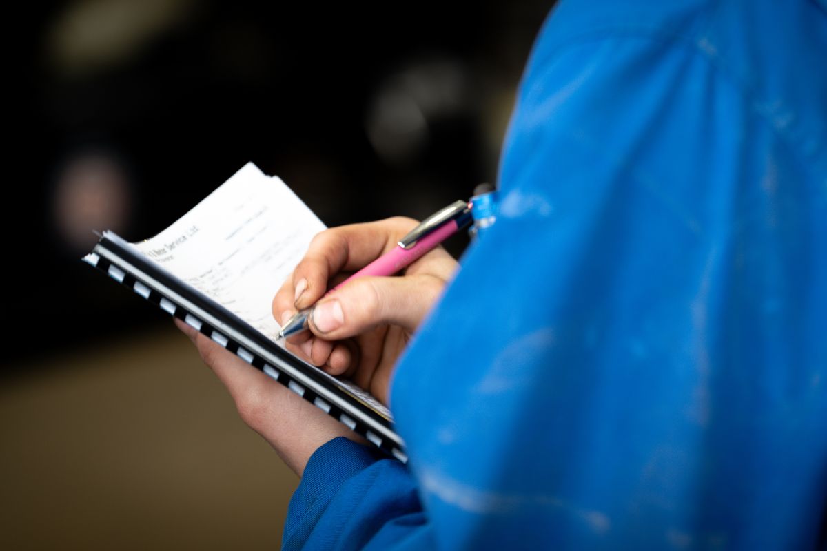 Person in a blue jacket writing in a notebook with a pen, focusing on note-taking during an activity or inspection.