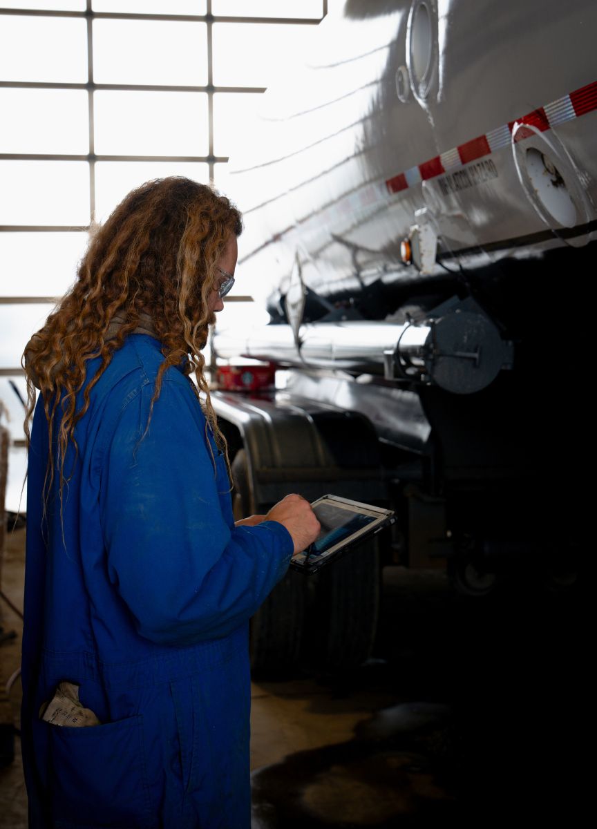 A Tru-Kare worker in blue coveralls using a tablet next to a large transport tanker  in the Tru-Kare shop.