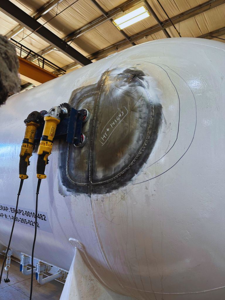 Close-up of a large, white spherical tank with a welded patch and marked lines for further repair, accompanied by two yellow grinders mounted on a magnetic drill stand, inside Tru-Kare, an industrial repair facility.