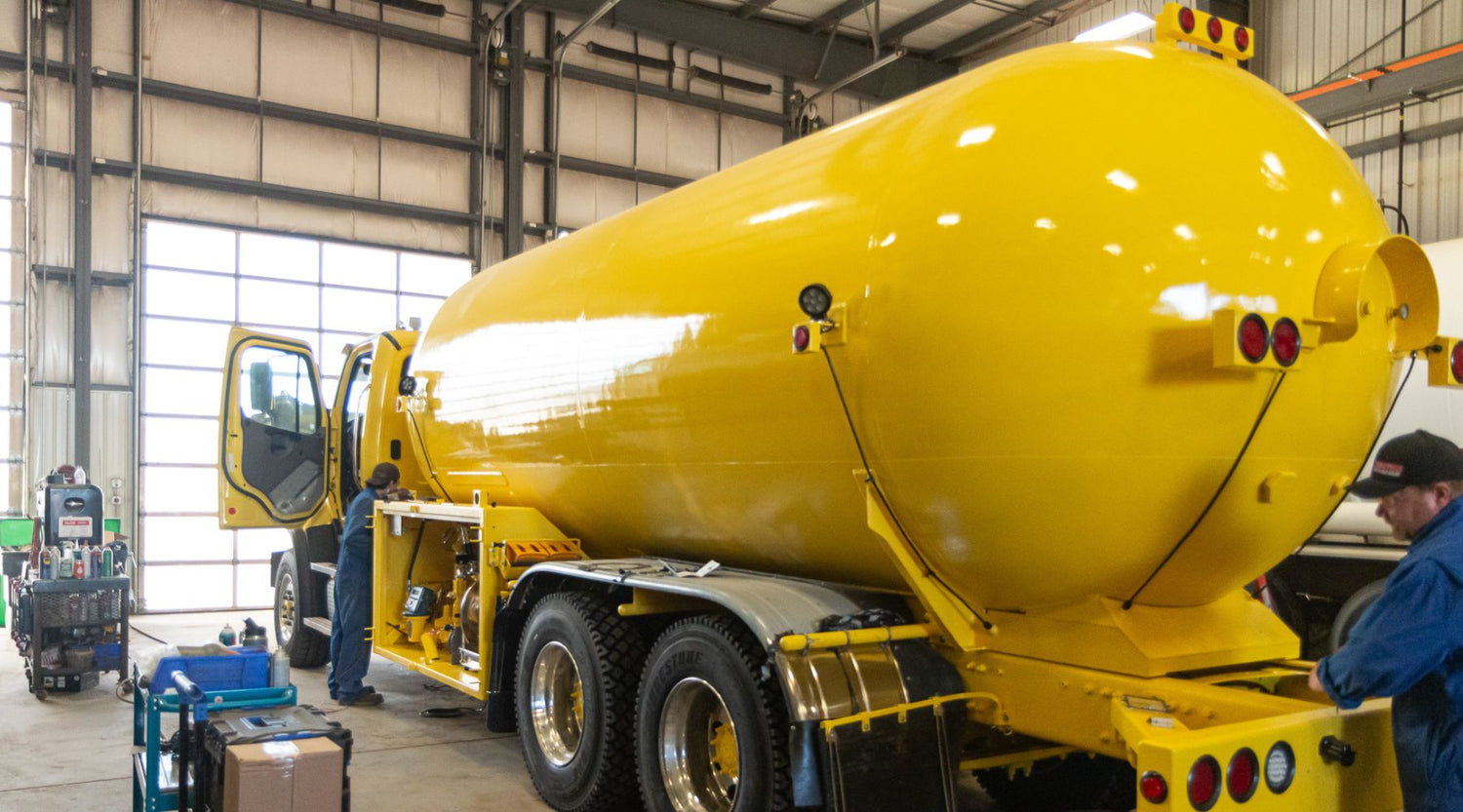 Workers inspecting and working on a large yellow industrial tank truck inside a workshop, with tools and equipment visible nearby.